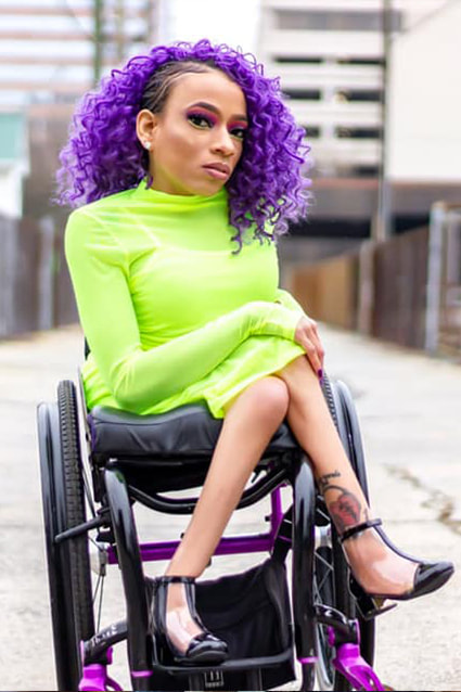 The image shows a Black woman in a wheelchair. The woman has black hair with a purple curly hair piece, and she is wearing a green long-sleeved dress. Additionally, she is wearing black shoes.