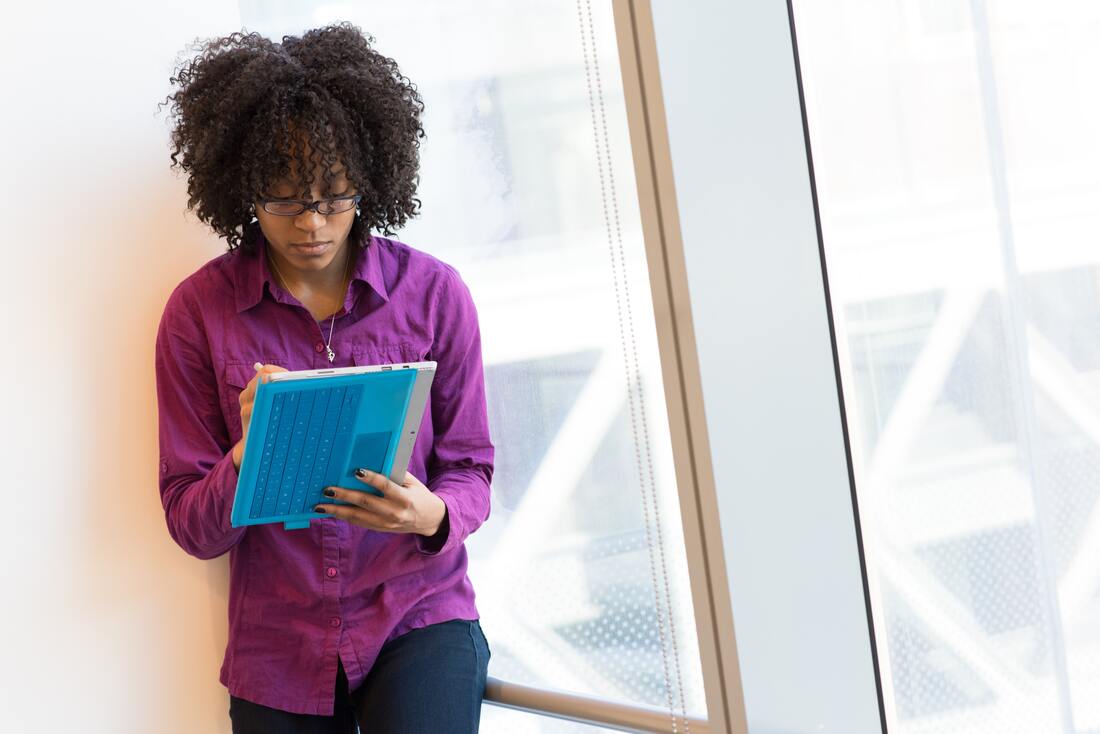 The image shows a Black woman standby a window and she is reading book. The woman is wearing purple blouse and blue trousers. Additionally, she has black curly hair and is. wearing glasses
