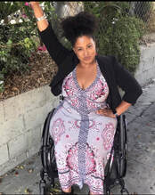 The image shows a Black woman, and she is in a wheelchair. She is wearing a purple dress. with a blue cardigan. Her black hair is tied up. She has one hand on knee and the other hand. raised in the air.